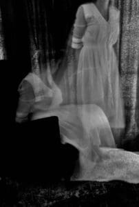 A haunting black and white photograph of a woman, captured with a long exposure effect that creates a ghostly appearance as she rises from a seated position. The ethereal quality of the image evokes the presence of maternal spirits, subtly hinting at the mystical realm of folklore and the enduring connections between the living and the departed.