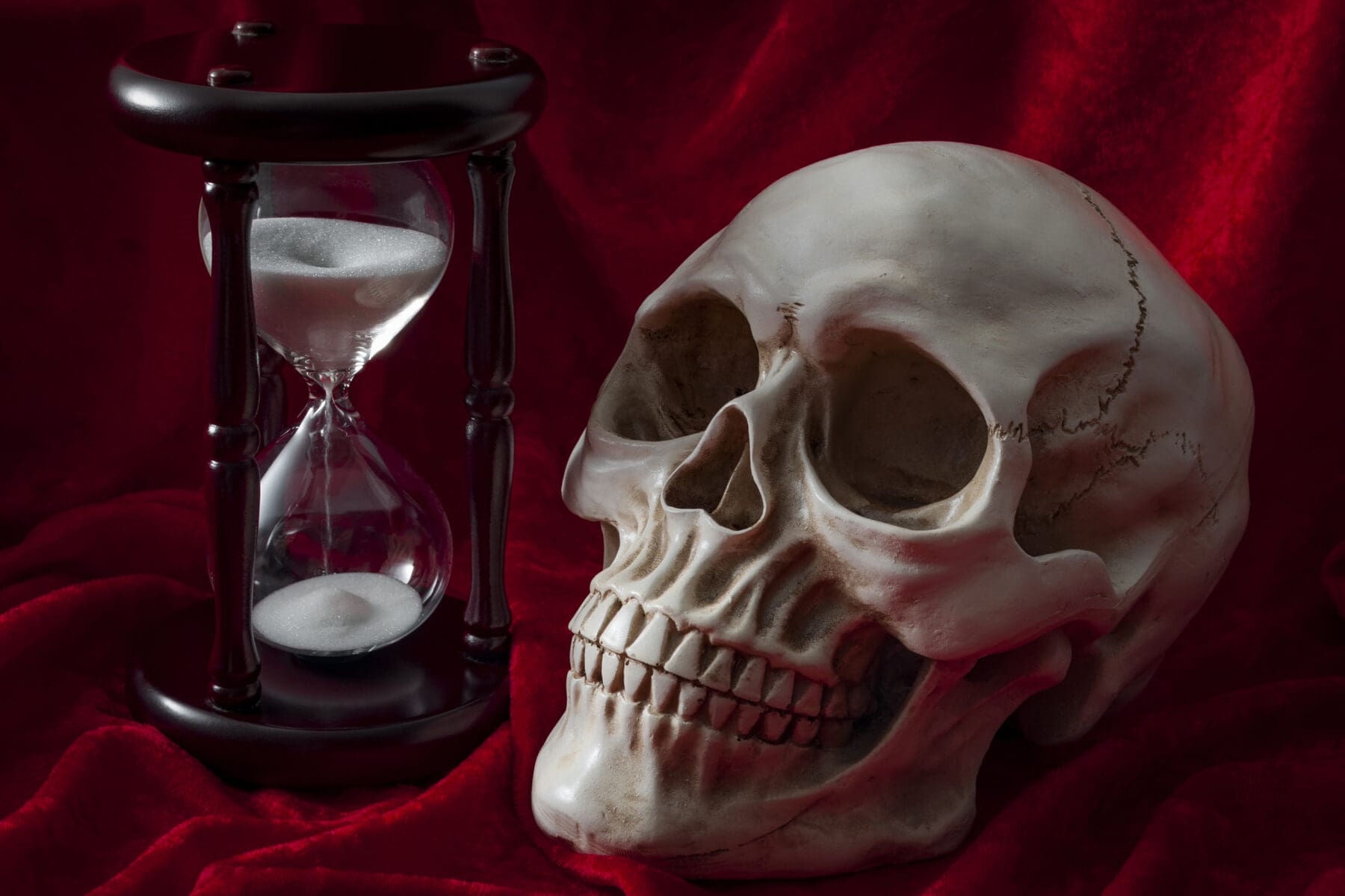 Ephemerality of life, fear of death and Memento Mori concept with a skull and vintage hourglass on red velvet background, dark tourism